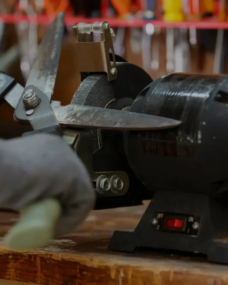 Hand i arbetshandske som slipar trädgårdsverktyg mot bänkslip i verkstad | Säffle Slipservice